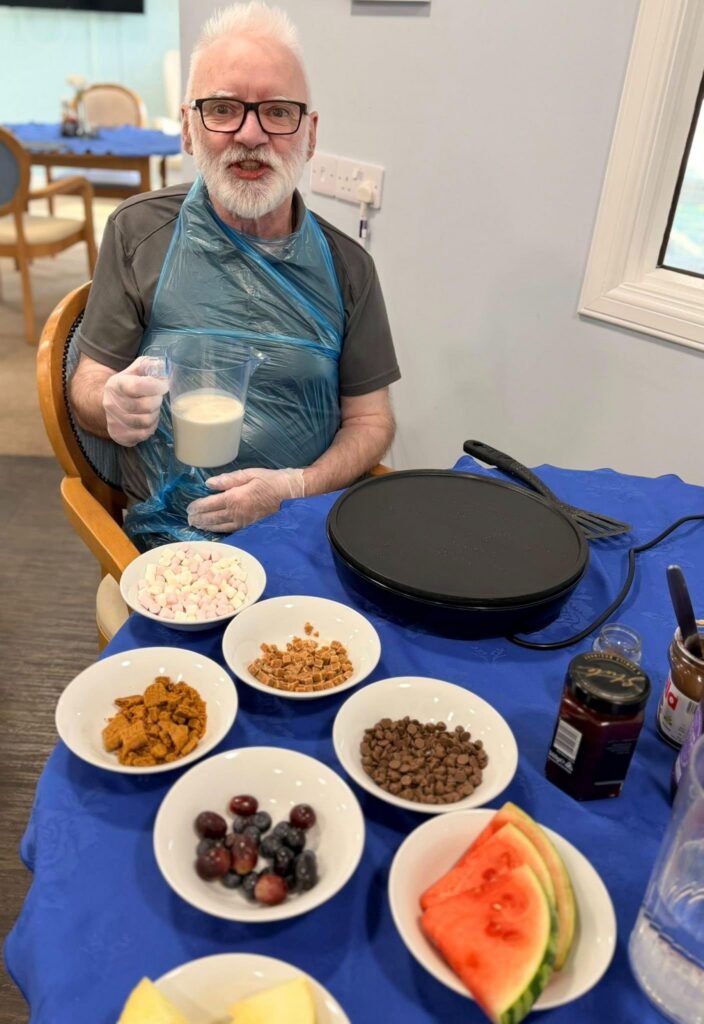 Man with jug of pancake batter smiling behind a grill and pancake toppings